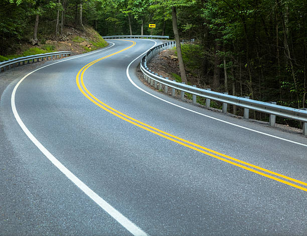 A road with freshly painted lines winds its way through the mountains of Pennsylvania. Guard rails and a directional sign help travelers stay on the road that is lined with trees on both sides.