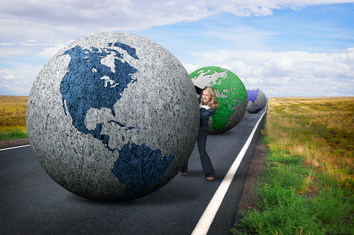 A businesswoman struggles to push a large globe down a long country road showing the Western hemisphere. She is being closely followed by two globes representing Europe, Africa and Asia as she leans out from behind the globe to look to see where she is going.