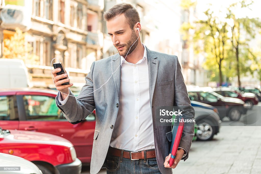 Businessman wearing earphone talking on smart phone on the street Young businessman standing on the street in the european city, talking on smart phone, wearing earphone. Headphones Stock Photo