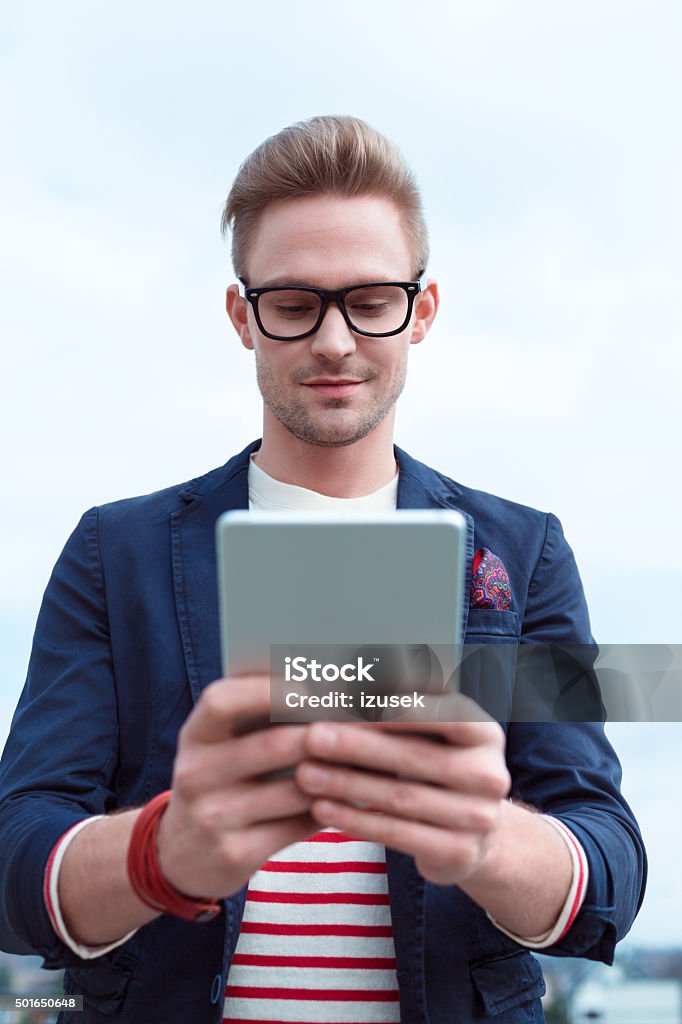 Blonde young man using a digital tablet outside Portrait of fashionable blonde young man wearing navy blue jacket, striped blouse and nerd glasses, using a digital tablet outdoor. Digital Tablet Stock Photo