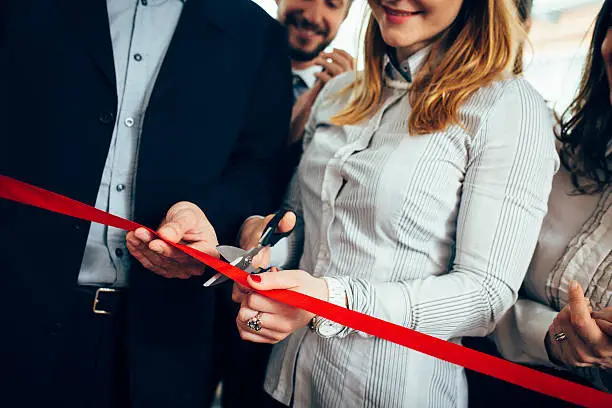 CEO and his assistant cutting a red ribbon, opening ceremony. Few people are in back and they are smiling. They are starting a new business.