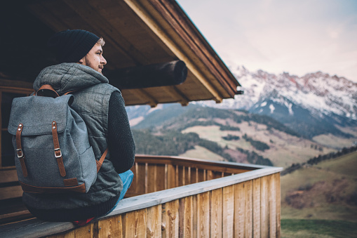 A man on vacation hiking in Austrian Alps. Sitting on the porch of wooden cabin. Wearing warm clothes and backpack. Preparing for long walk and enjoy in beautiful landscape. Winter day. Mountains are covered with snow.