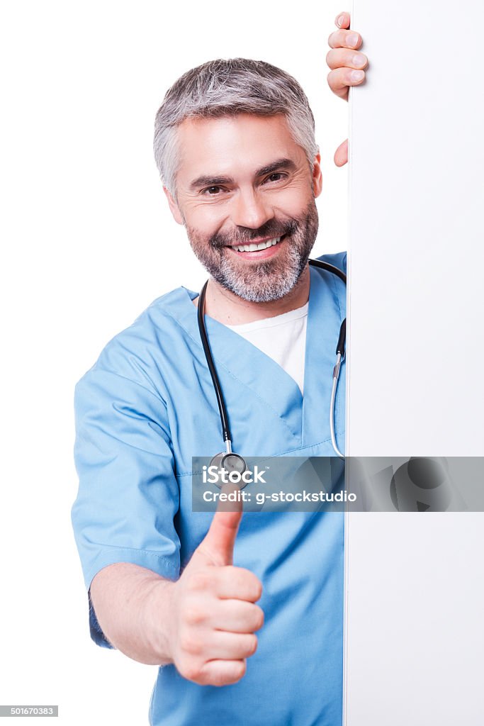 Successful surgeon. Confident mature surgeon looking out of the white poster and showing his thumb up while standing isolated on white Active Seniors Stock Photo