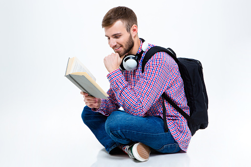 Cheerful handsome bearded young man in checkered shirt with backpack and headphones reading book and sitting with crossed legs