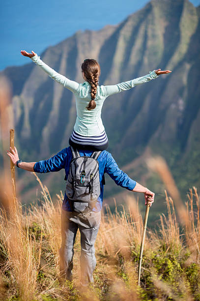 A woman is sitting on her husbands shoulders on the top of a Hawaiian mountain.