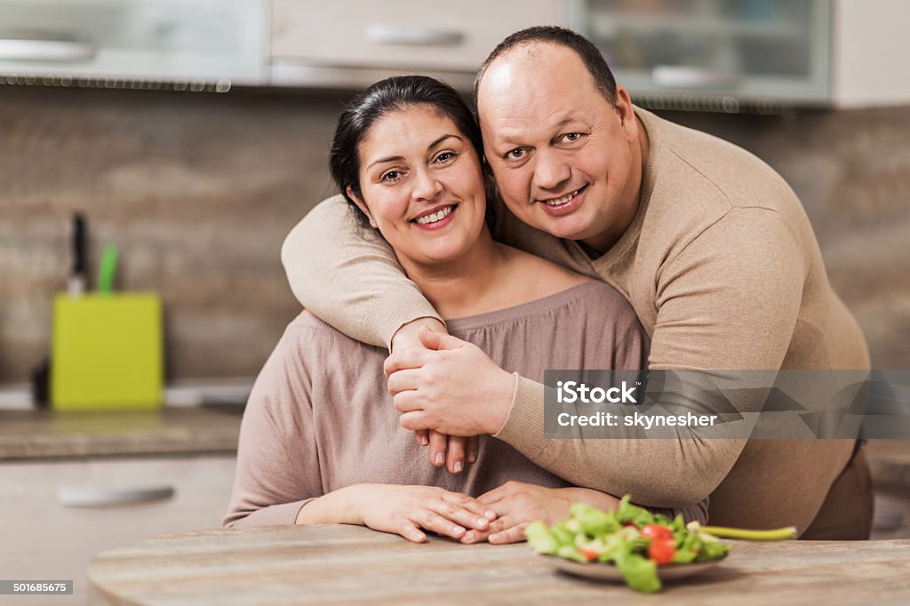 Happy overweight couple. Happy overweight embraced couple in the kitchen looking at the camera.    Men Stock Photo