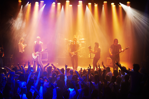 Musician playing electric guitar onstage at a music venue, with a drummer and a videographer in the background.