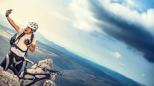 Young adult mountain biker with a helmet and a sophisticated black mountain bike smiling and posing for a selfie on top of a mountain. Green valley with forests in the background.