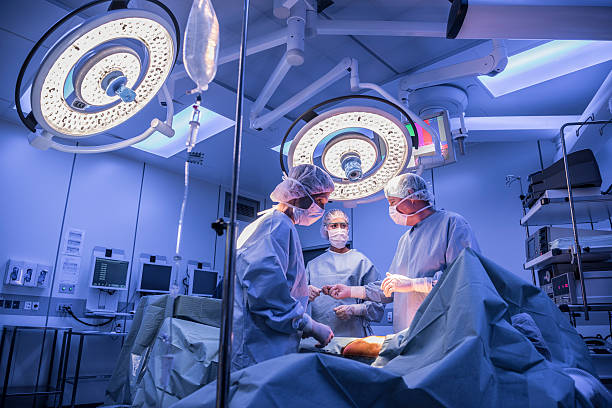Surgeons operating on patient in operating theatre under lights Doctors wearing surgical masks and gowns performing an operation on patient in hospital operating theater. surgeon stock pictures, royalty-free photos & images