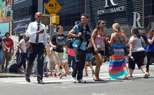 New York, NY, USA - July 2, 2014: Businessmen with smart phones and tourists with shopping bags among people crossing a busy intersection along 7th Avenue in New York City.