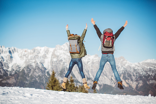 Couple on vacation hiking with a backpack in Austrian Alps. Wearing warm clothes. Jumping on the top and enjoying in beautiful landscape. Winter day. Mountains are covered with snow.