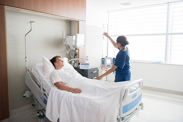 Female nurse tending to male patient in hospital bed Young man in hospital being cared for by nurse. Medical professional looking after patient on hospital ward. Adjusting iv line, medication, care, bedside manner. hospital patient bed nurse stock pictures, royalty-free photos & images