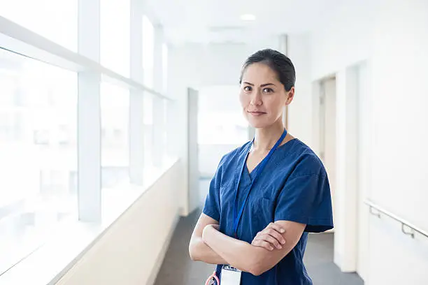 Female nurse looking towards camera, wearing blue scrubs, arms crossed. Medical professional working in hospital. Attractive female doctor at work.