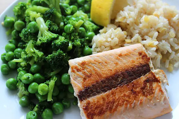 Photo showing a healthy meal consisting of a dry fried salmon fillet served with brown rice and red lentils, garden peas / frozen peas mixed with small pieces of broccoli and a slice of lemon.  This dish is part of a healthy eating, low calorie diet plan.