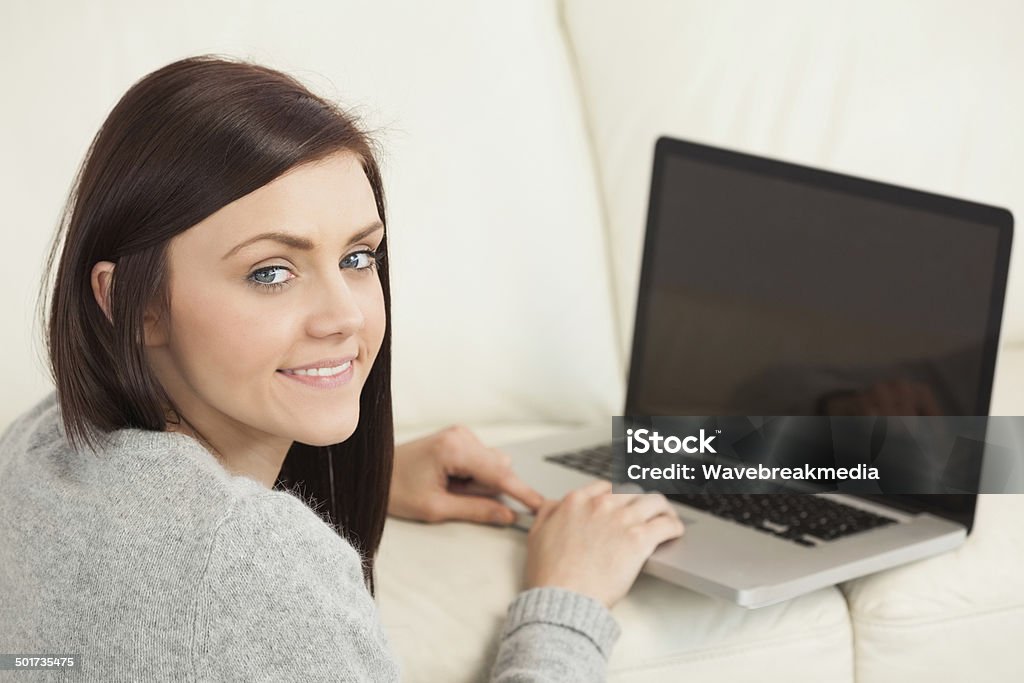 Garota sorridente usando um laptop no sofá olhando para a câmera - Foto de stock de Aconchegante royalty-free