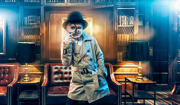 Boy detective wears a typical old school detective  trench coat and a fedora hat. He stands inside a living room with old furniture and book shelves. The detective boy holds a magnifying glass in front of one eye as he searches for clues.