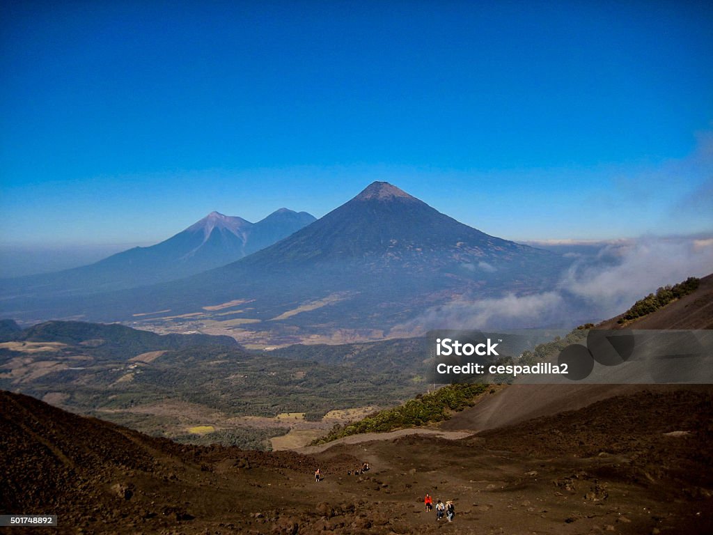 Volcanoes in Guatemala, as seen from the Pacaya Volcano. This photograph shows 4 volcanos in Guatemala, the Pacaya Volcano (being climbed), the Agua Volcano, Fuego Volcano (active and with the top charred) and the Acatenango Volcano. Guatemala has a few active volcanos, and many more dormant ones, and they help shape the landscape of the country. In the photograph, these colossal landmarks rise agains the blue sky, creating an impressive, beautiful view. 2015 Stock Photo