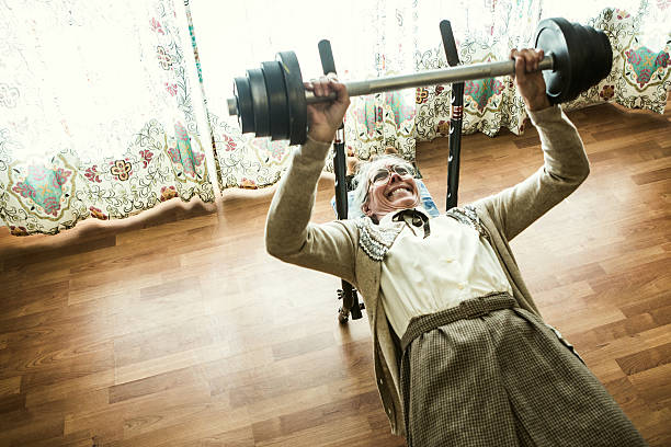 Grandma Weightlifting in Living Room A senior woman performs a bench press with a barbell loaded with weights in her living room, wearing her normal skirt and sweater.  Horizontal image with copy space. senior bodybuilders stock pictures, royalty-free photos & images