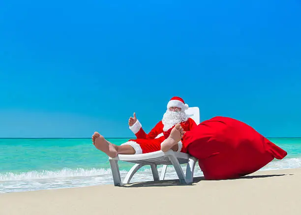 Christmas Santa Claus with big red sack thumbs up gesture by hand, relax on deckchair at ocean tropical beach - New Year travel destinations for vacation in hot countries concept