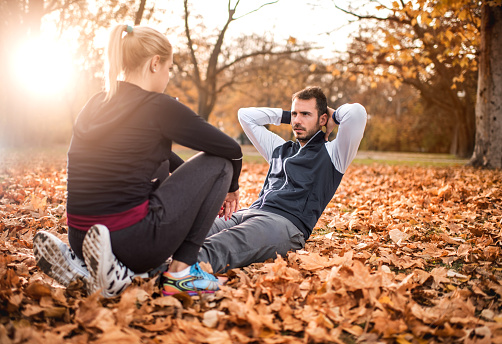 Athletic man exercising sit-ups with a help of his female friend during autumn day in nature.