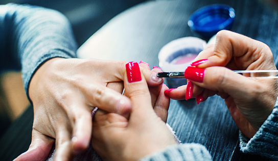 Closeup of unrecognizable woman having her fingernails covered with translucent gel before red nail polish is being applied by professional manicurist at nail salon. Back lit, toned image.