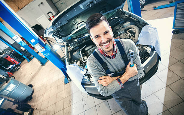 Car mechanic proudly posing. Wide angle portrait of smiling mid 30's car mechanic standing in front of a vehicle he's attending to. Holding a wrench in his hand. Top view. car mechanic top view stock pictures, royalty-free photos & images