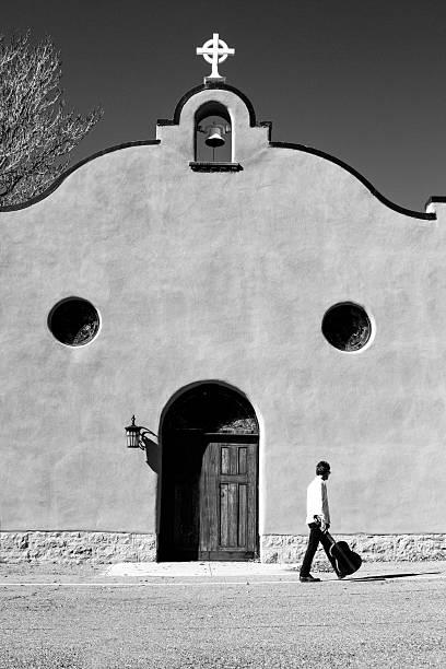 A B&W shot of a man walking with his guitar in front of a Southwest US adobe church. Shot in Santa Fe, NM at the Rosario Chapel, also known as the Church of Our Lady of the Rosary.