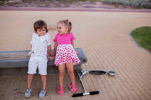 Cute brother and sister relaxing on a bench while girl is saying something to her little brother.