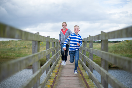 Two brothers running across a bridge over water in the countryside. They are wearing casual clothing and smiling.