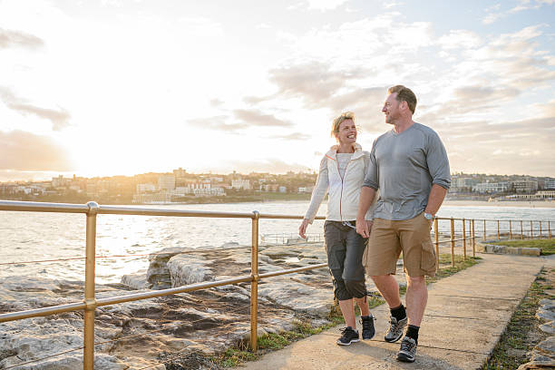 Mature couple walking on boardwalk at sunset, Bondi Beach Couple walking by sea on path on vacation in Sydney, Australia. boardwalk stock pictures, royalty-free photos & images