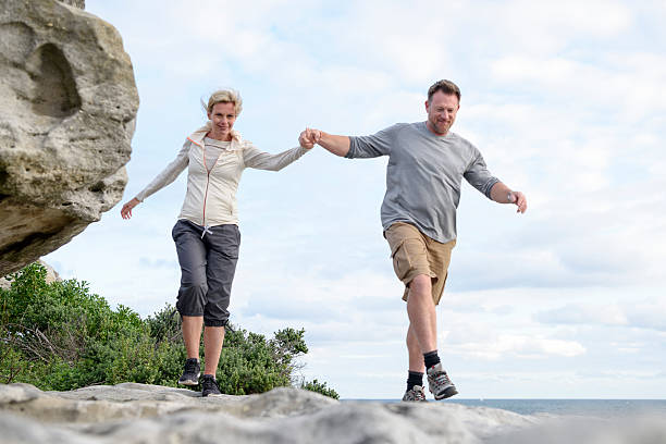 pareja madura excursionismo en rocks holding hands, playa de bondi - couple mature adult action walking fotografías e imágenes de stock