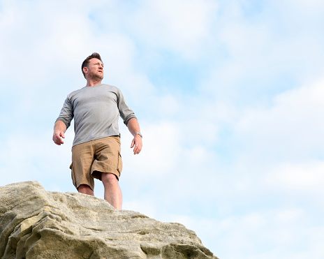 Male hiker looking at view from top of rocks, view from below. Man on hiking vacation against a clear sky.