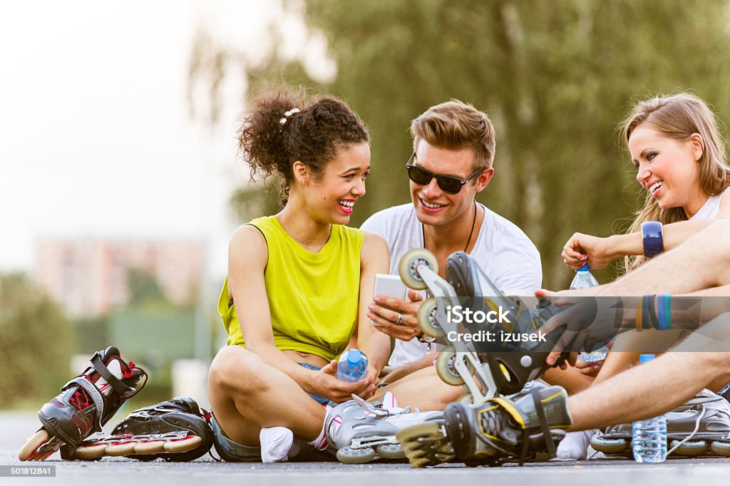 Young people on rollerblades Young people resting after rollerblading, sitting on the tarmac and talking. The boy holding smart phone in hand. Skate - Sports Footwear Stock Photo
