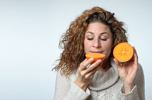 Healthy young woman enjoying a halved fresh ripe juicy persimmon smiling with anticipation as she prepares to take a mouthful while displaying the second half to the camera