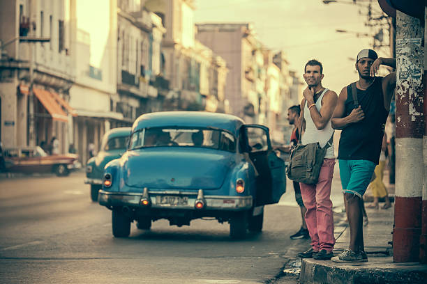 Havana, Cuba-November 7, 2015. Busy residential street of Centro Havana. Two young men (one white, one black), waiting on the street corner for a taxi. Other people int he background. Old, rundown residential buildings, remembering better times from 50 years ago. Street life in full effect: lot of people on the street. Facades are in bad shape due to long time neglect and poverty. Traffic in the background.