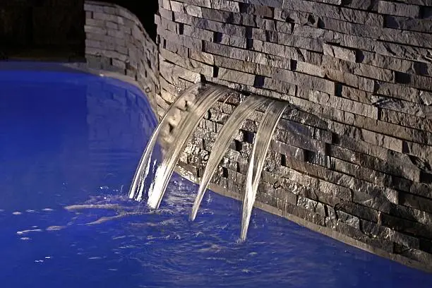 A night-time study of the tranquility of a poolside water-feature forming a small laminar waterfall. The feature is set into a modern-style masonry wall. The pool lighting resulted in the unusual deep-blue color of the water. 