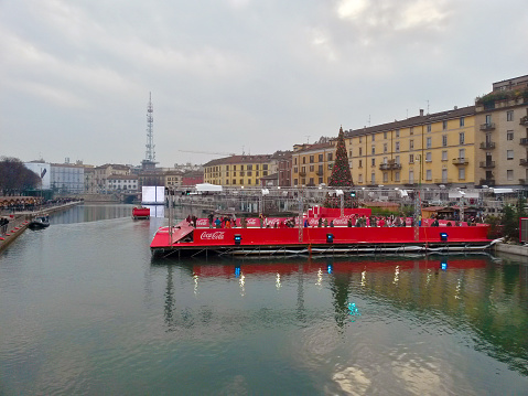 Milano, Italy - December 13, 2015:  Christmas village on Darsena. Skating ring sponsored by Cocacola with some people who are skating. Many people walking on promenade. One electronic screen on background.