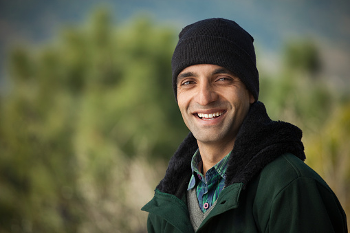 Outdoor image in winter season of a happy young man standing in fresh air and giving toothy smile while looking at camera. The man is wearing winter clothes, jacket, cap and shirt. One person, hand and shoulder and horizontal composition with copy space.