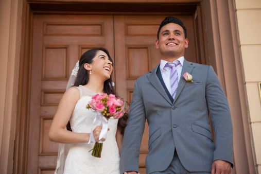 A radiant couple in wedding attire laugh and hold hands against the grand columns of a city building, their joy palpable in the golden sunlight.
