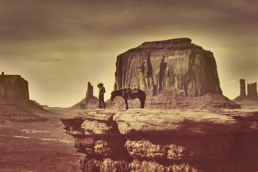 Retro styled, sepia toned, old-fashioned photograph of a Native American Indian cowboy standing with his horse on a rocky cliff and looking into the Southwest USA desert wilderness of Monument Valley Tribal Park, Arizona. Navaho tribe man with saddle animal admiring Wild West extreme terrain beauty of the Indian Reservation landscape.