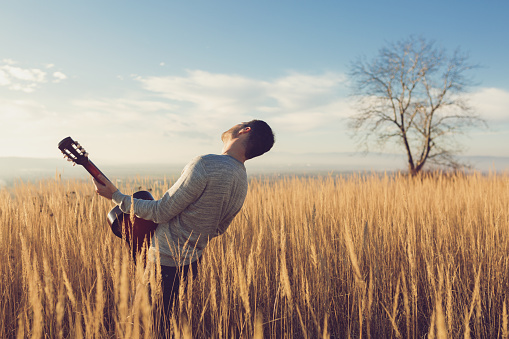 Man is playing a guitar while standing in the middle of a prairie, enjoying a beautiful sunny day in the nature far away from the city life.