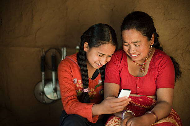 Indoor image of Asian daughter and mother sharing mobile phone. Indoor image shot in a small adobe kitchen of peasant family where a happy daughter showing mobile phone to her mother. They are clad in traditional Hindu dress. Two people, three quarter length, horizontal composition with selective focus and copy space. nepal photos stock pictures, royalty-free photos & images