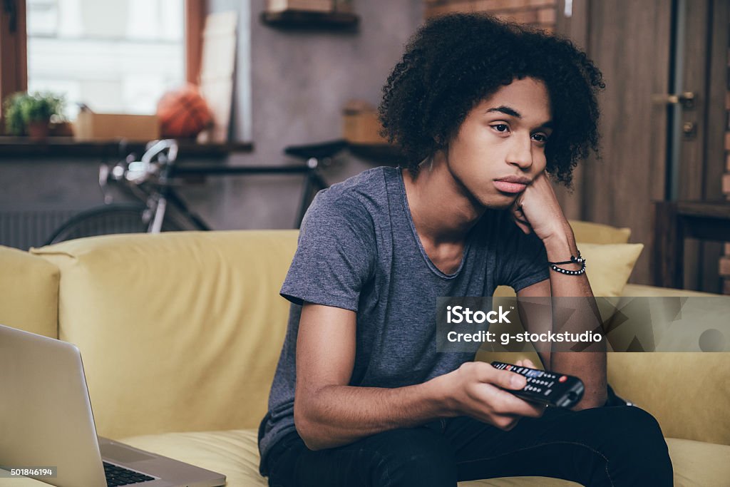 Nothing interesting to watch. Young African man holding remote control and looking bored while watching TV on the couch at home Boredom Stock Photo