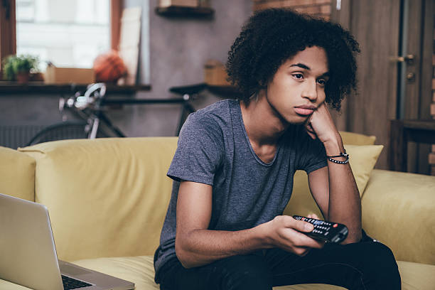 Nothing interesting to watch. Young African man holding remote control and looking bored while watching TV on the couch at home boredom stock pictures, royalty-free photos & images