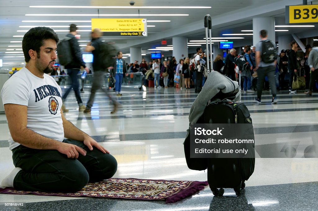 Muslim Praying in Airport New York, United States - December 17, 2015: Muslim Praying in Airport creating within himself his spirituality moment not regarding about the airport caos that there is around him. A long gate cue is present at the gate but nevertheless the muslim is fully concentrated in his daily praying. The muslim is male of about 30 year old wearing a white t-shirt.  Islam Stock Photo