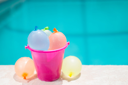Pink bucket filled with water balloons poolside.