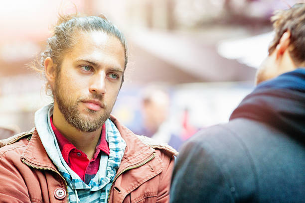 Young man with a scruffy look listens to his friend with a serious expression. They are both standing in the street.