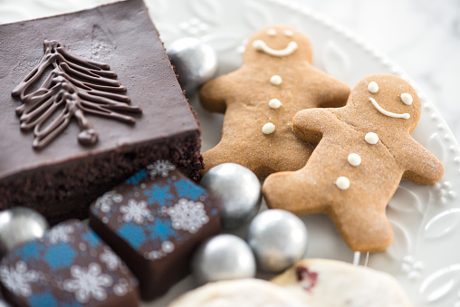 A welcome plate of Christmas desserts at a luxury hotel.  A modern selection and assortment of seasonal Xmas treats on a white country styled plate with holly leaves including a chocolate brownie with tree drawn in icing, silver cola ball candies, luxury snowflake decorated chocolates, gingerbread man cookies and cranberry shortbread.  Set on a marble table.  Blue, white, silver, brown colours.