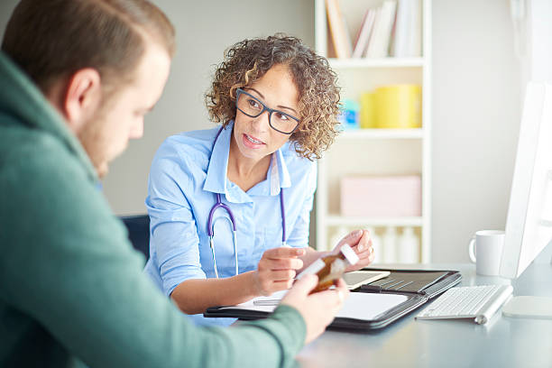 female gp precription advice A female doctor sits at her desk and chats to a male patient about his current medication . She is dressed in a shirt with rolled up sleeves . They are both looking down at the pill bottle as she assesses his current dosage . prescription medicine stock pictures, royalty-free photos & images