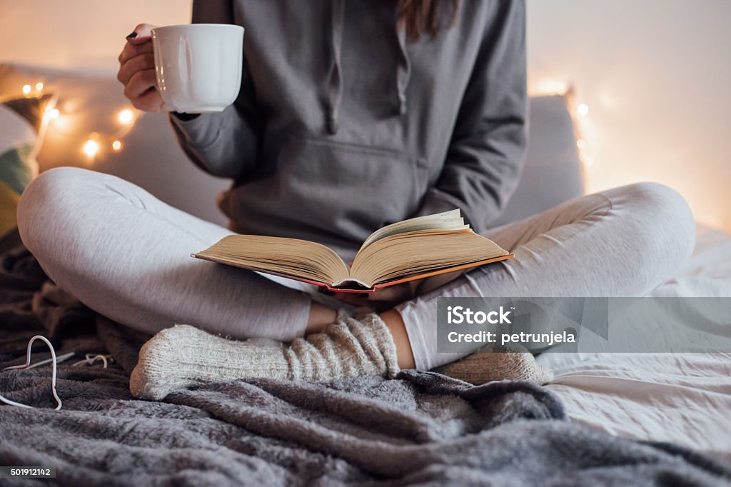 Girl drinking hot tea and reading book in bed Girl holding cup of hot tea and reading in bed. Around her in bad earphones, book, smart phone. Decorative lights in background. Reading Stock Photo